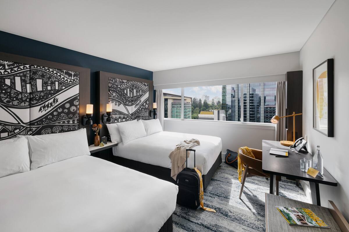 A hotel room with two beds, a desk, a chair, a suitcase, and a view of the city.