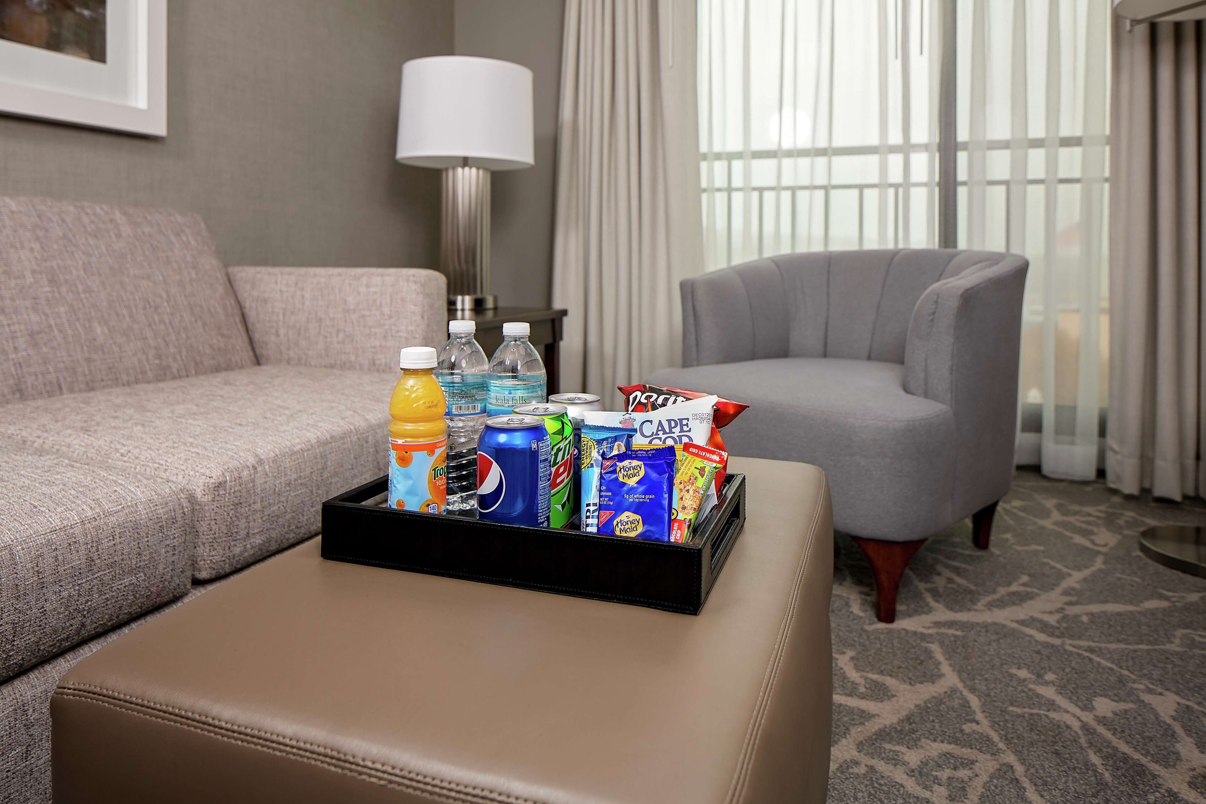 Habitación de hotel con sofá gris, un sillón gris, una mesita con una bandeja de aperitivos y bebidas, y una lámpara.
