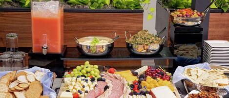 Un bufé con diversos alimentos, incluyendo frutas, quesos, carnes y un dispensador de bebidas.