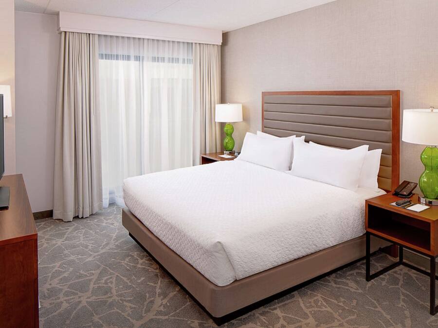 A hotel room with a large bed, two bedside tables with lamps, a TV on a stand, and a window with curtains.