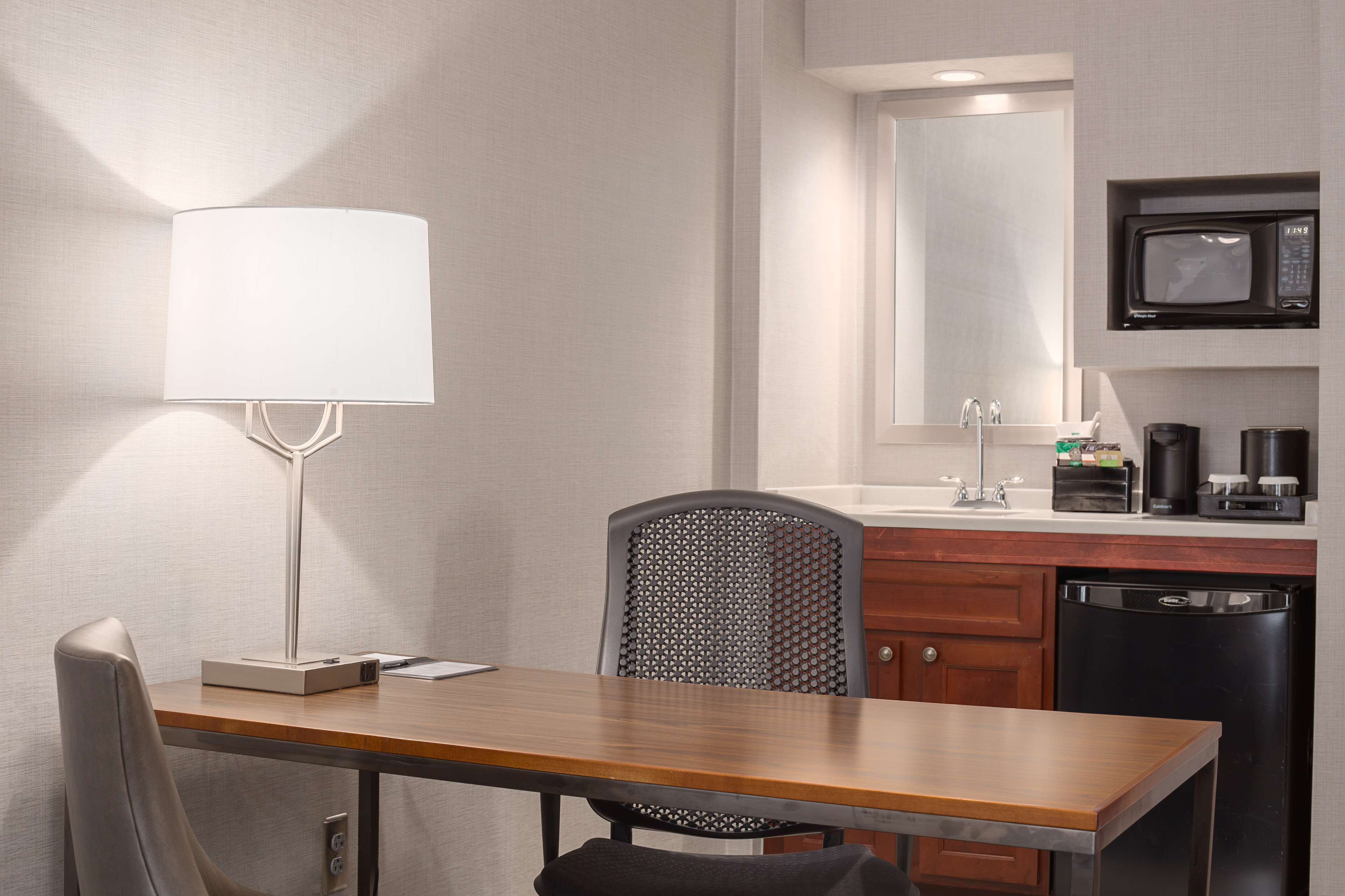 Habitación de hotel con escritorio de madera, una silla, lámpara, microondas, cafetera y lavavajillas.