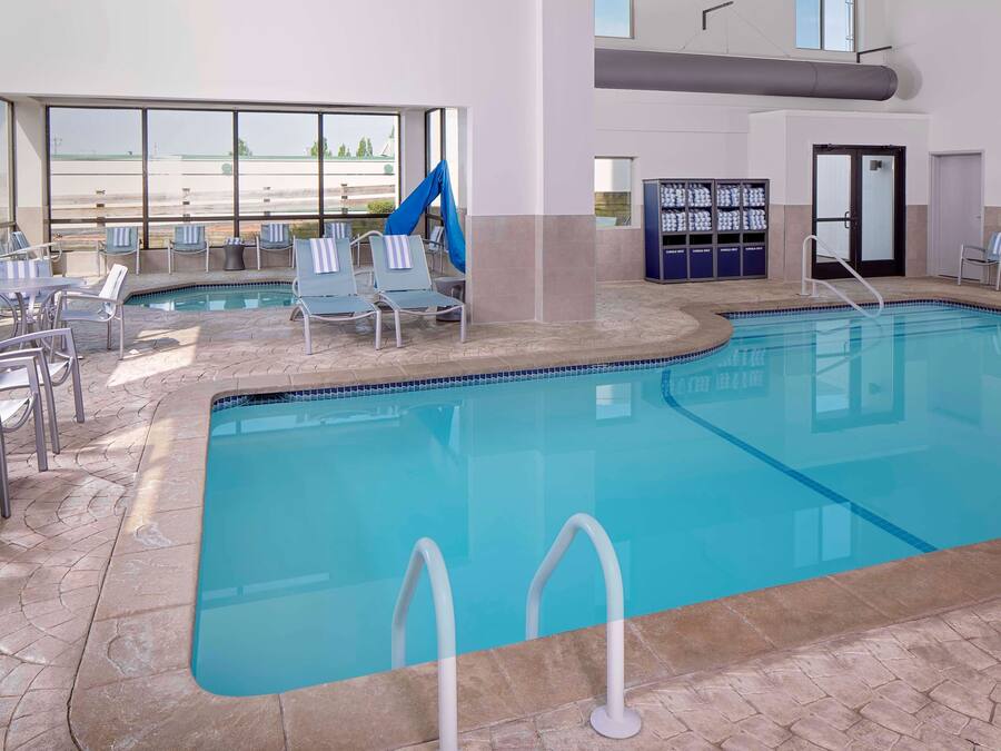 An indoor swimming pool with lounge chairs and a slide, surrounded by large windows and a tiled floor.