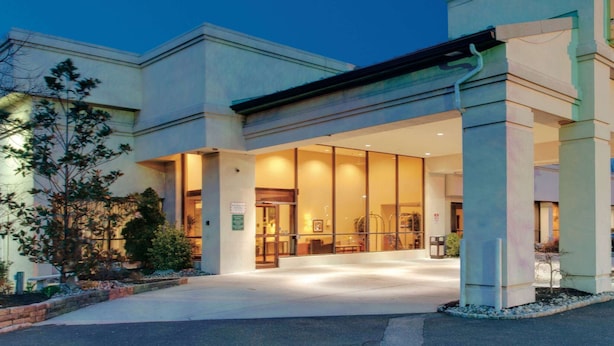 Entrada moderna de hotel com fachada de vidro, área de acesso coberta e jardim ajardinado.