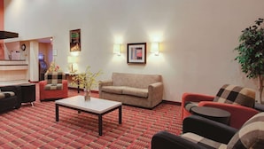 Um hall de hotel com carpete estampado em vermelho, um sofá xadrez, uma poltrona vermelha, uma poltrona preta, uma mesa pequena com um vaso e duas luminárias fixadas na parede.