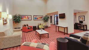 Um hall de hotel com carpete vermelho, várias áreas de estar incluindo sofás e poltronas, duas plantas em vasos, uma TV de tela plana na parede e uma mesa com um computador.