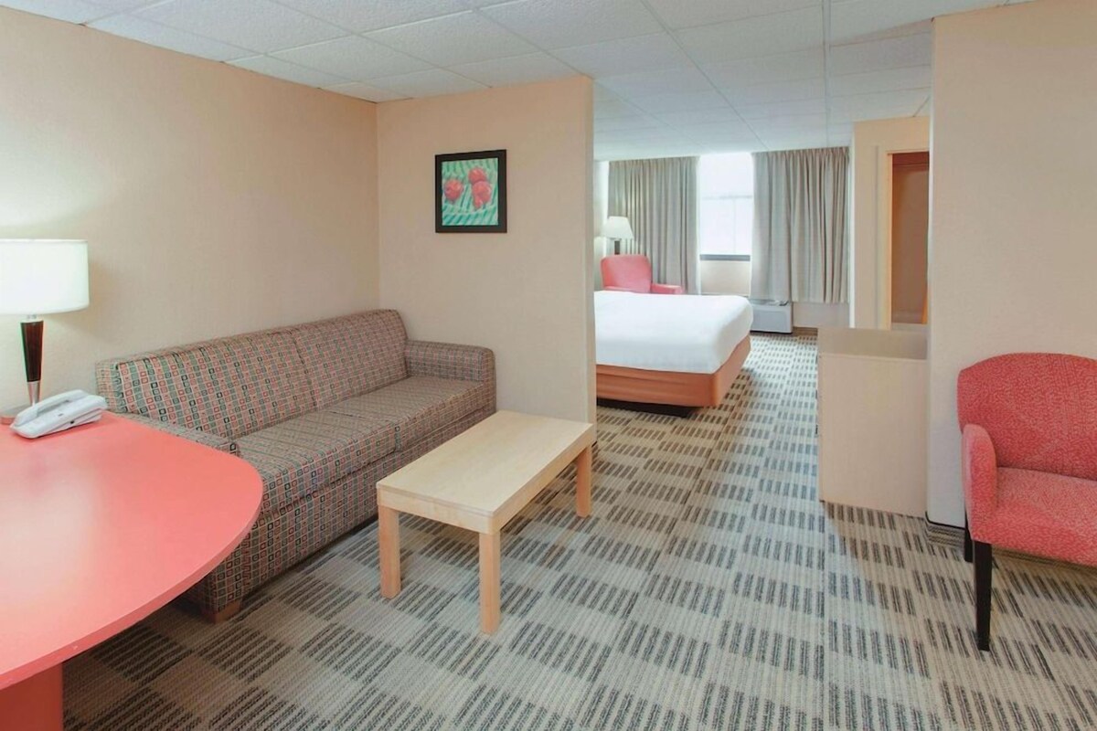 A hotel room with a patterned sofa, a small table, a bed, and a red chair.