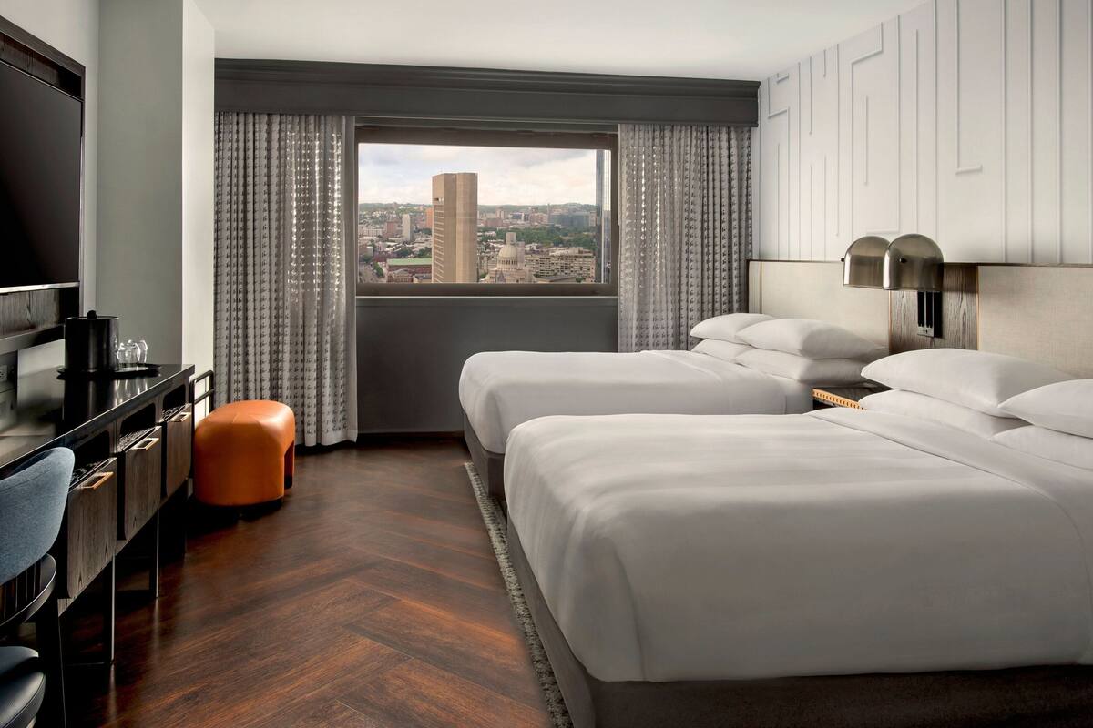 A hotel room with two beds, a desk, a chair, and a view of the cityscape.