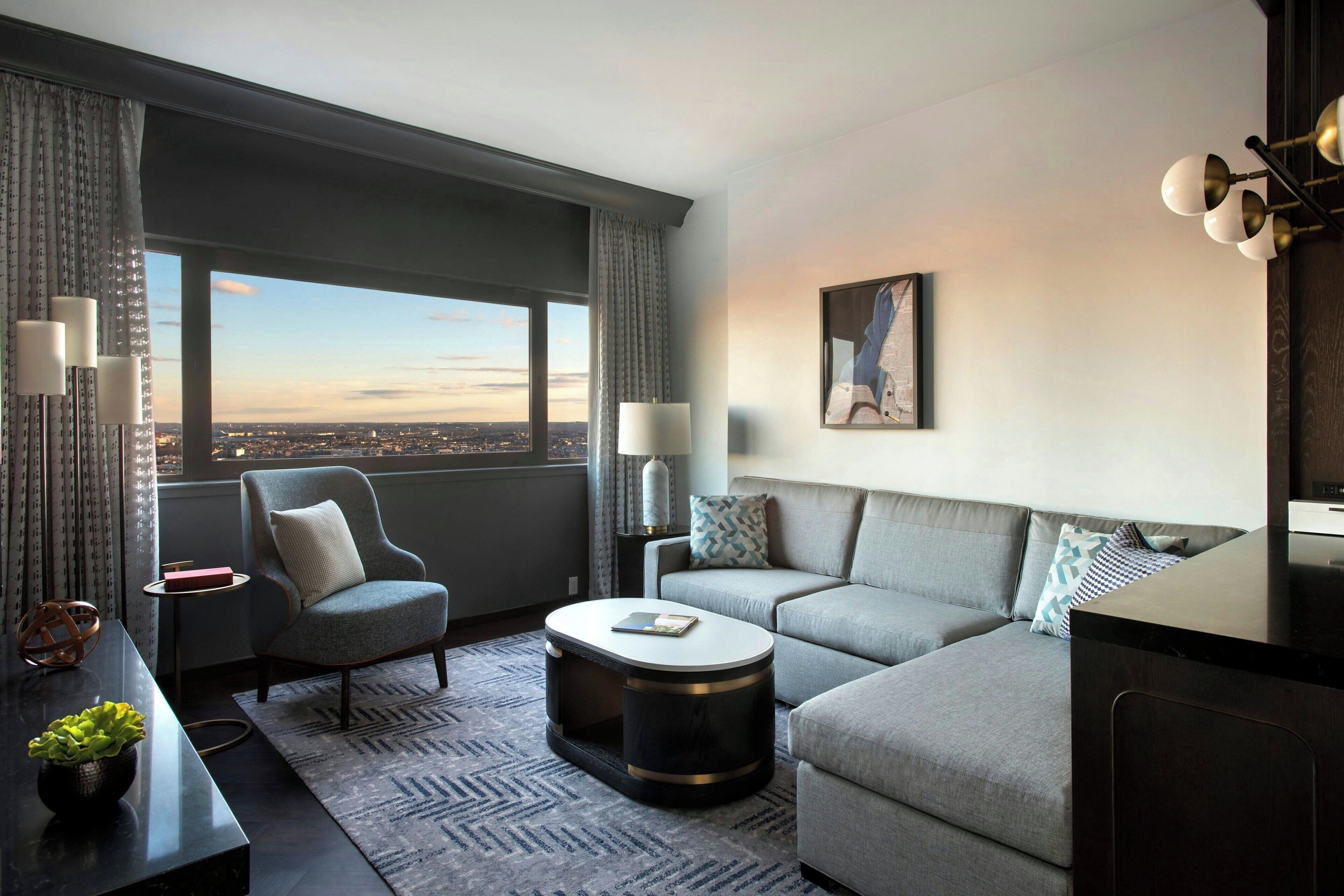 A modern hotel room with a large window, a sofa, a chair, a coffee table, and a lamp.
