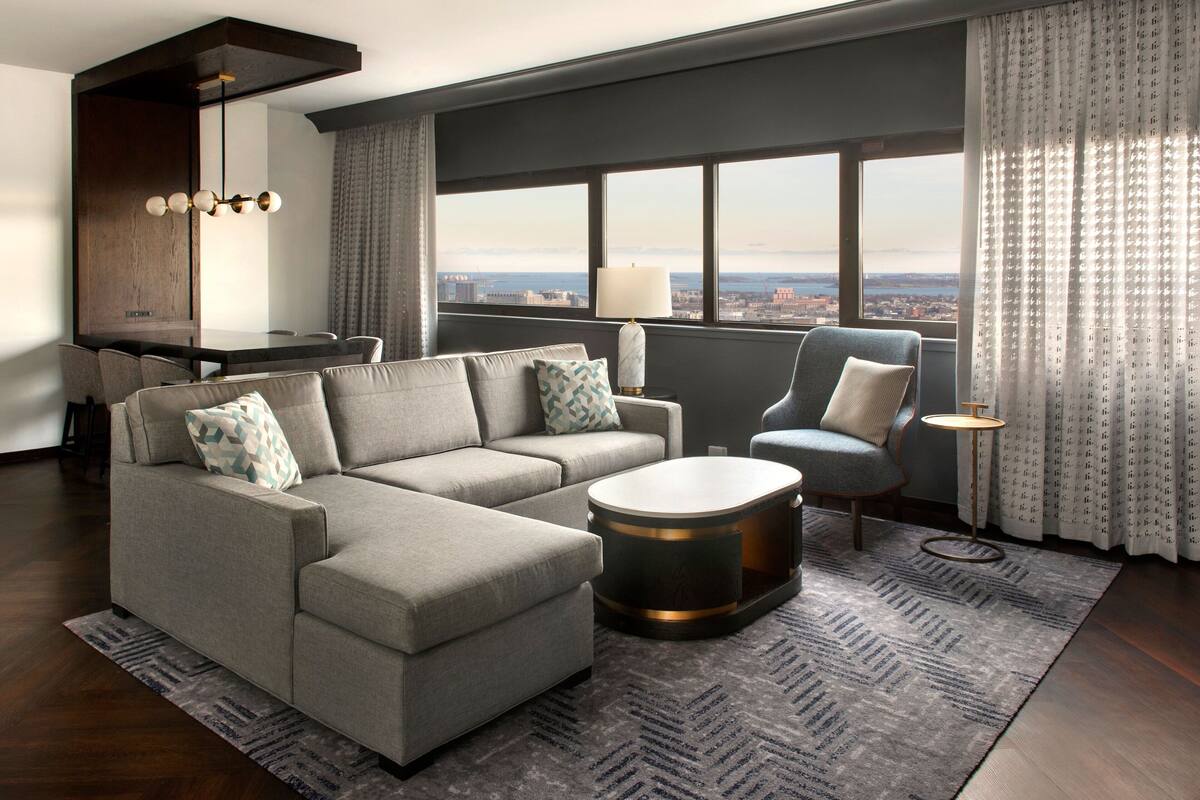 A modern living room with a gray sectional sofa, a round coffee table, and a blue armchair. There is a dining area with a dark wood table and chairs, and a view of the city through large windows.