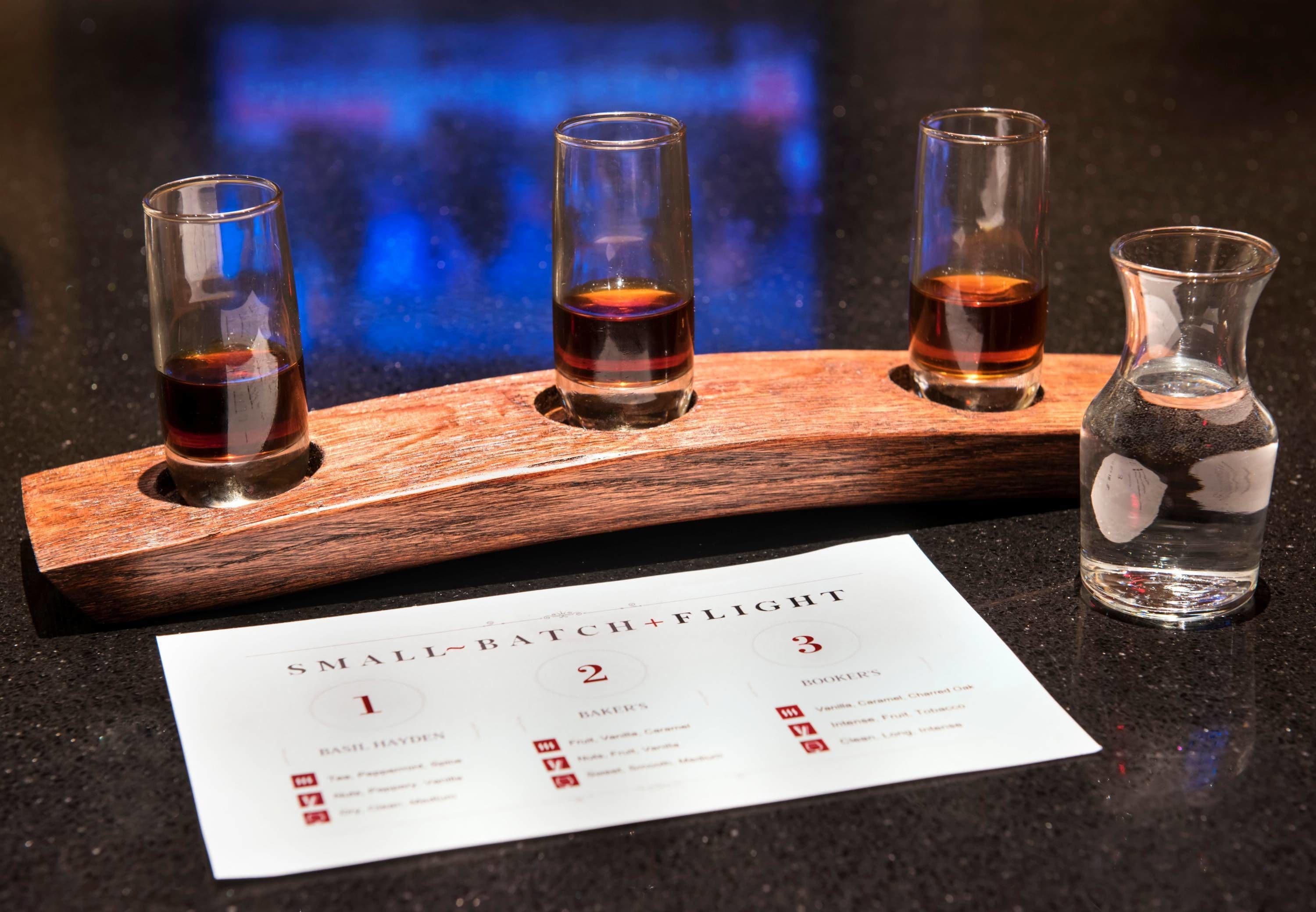 A wooden tray holding four shot glasses with a menu card and a bottle of water on a dark countertop.