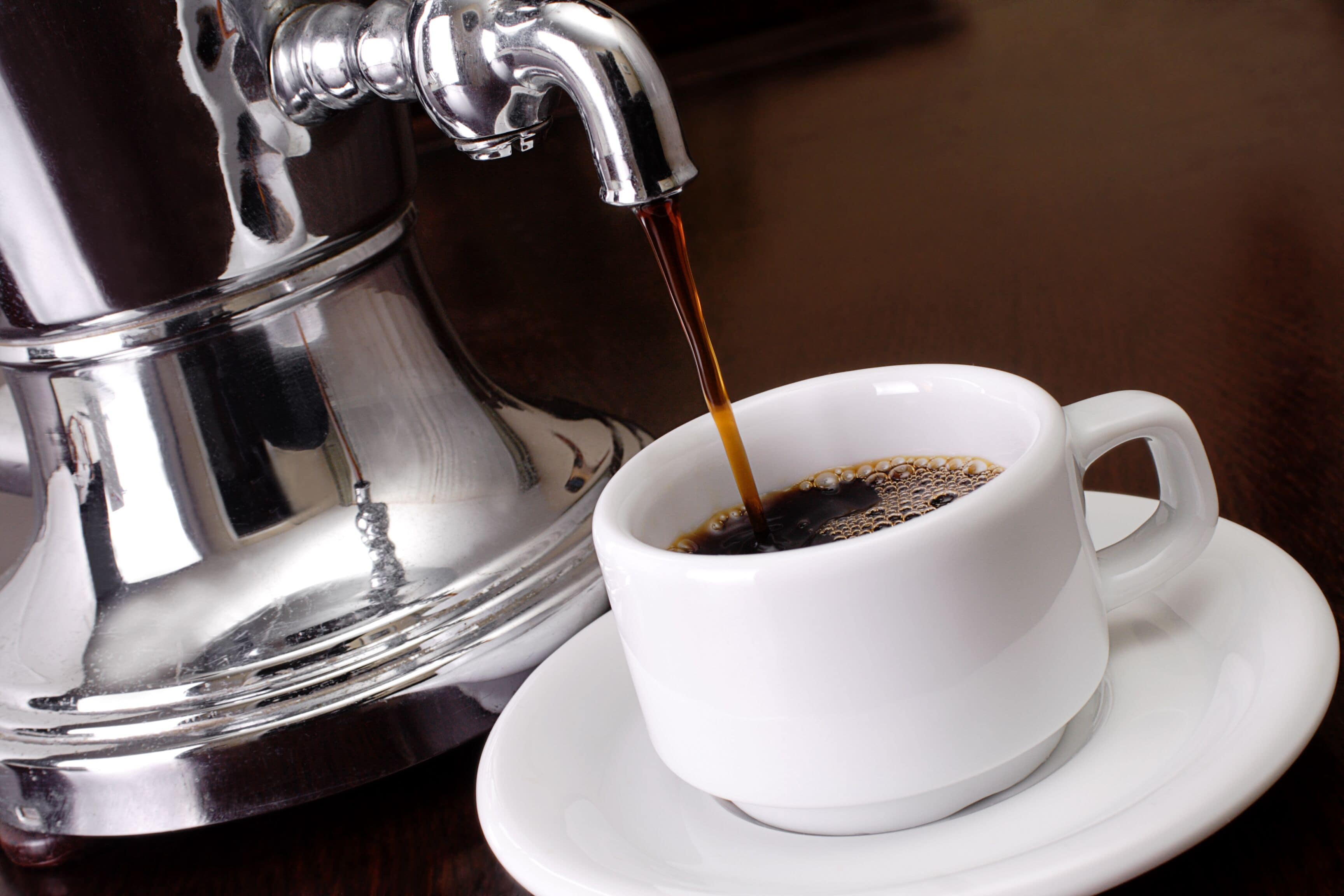 Una cafetera vertiendo café en una taza blanca sobre un platillo.