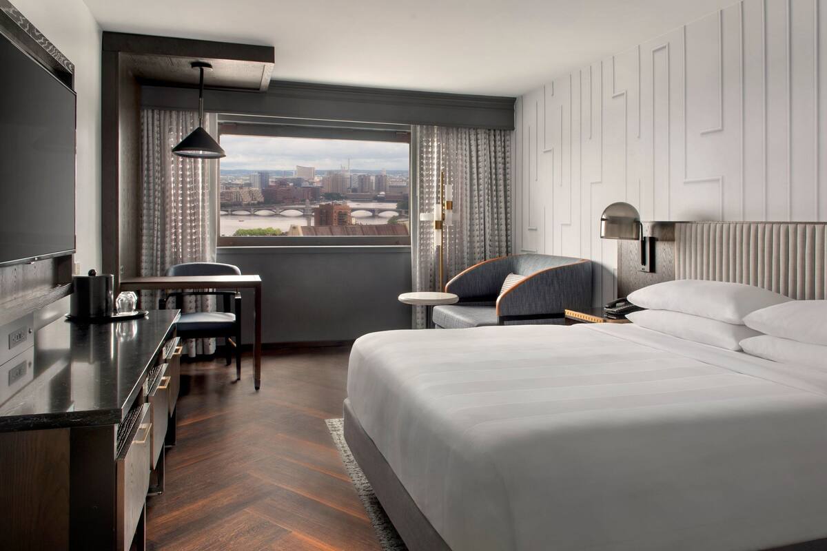 A modern hotel room with a large bed, a desk, a chair, and a view of the city through a large window.
