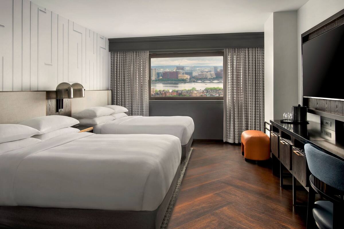 A hotel room with two beds, a desk with a TV, and a view of a cityscape.