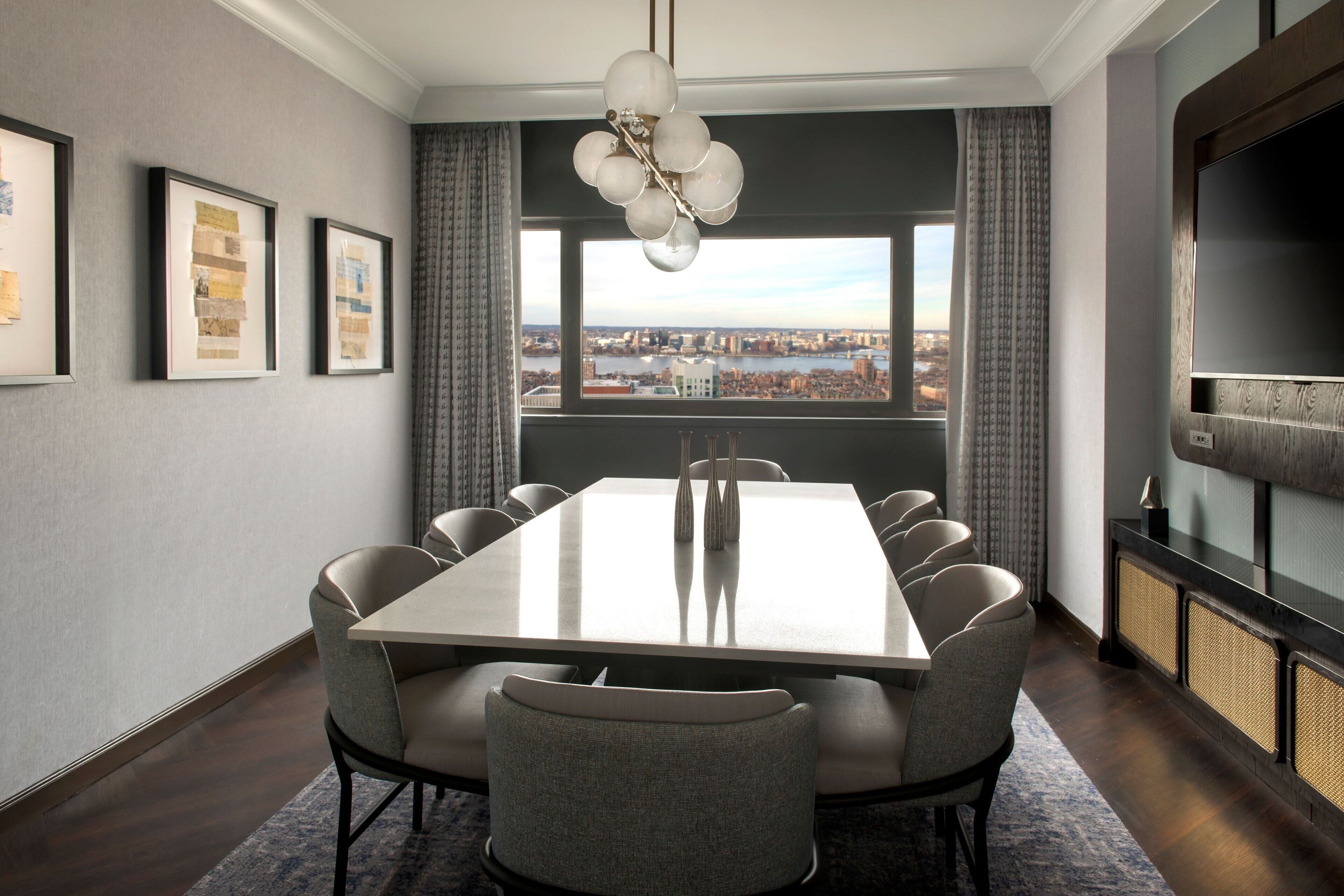 A modern dining room with a large table, comfortable chairs, a chandelier, framed artwork on the wall, and a city view through the window.