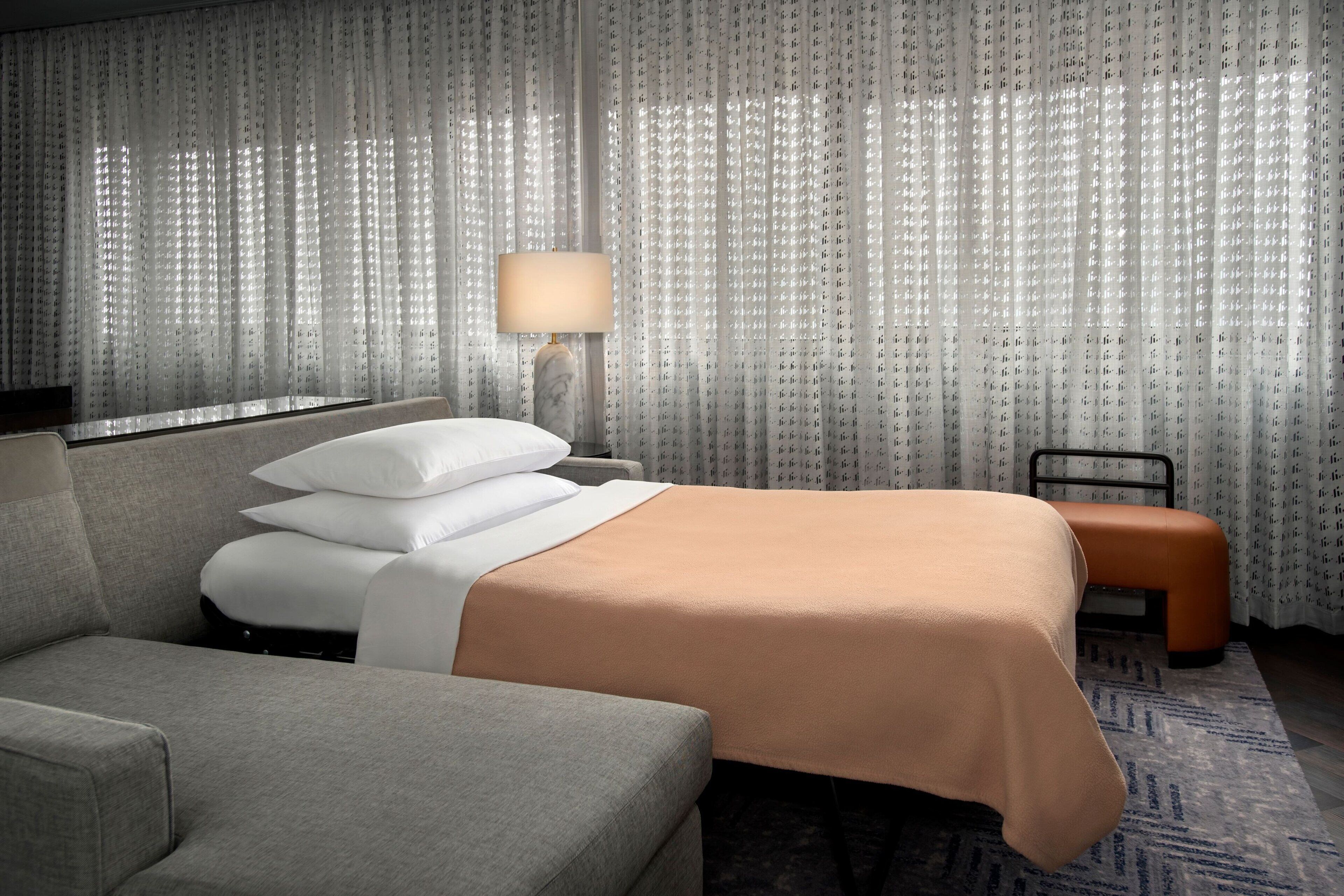 A hotel room with a bed, a sofa, a nightstand with a lamp, and sheer curtains.
