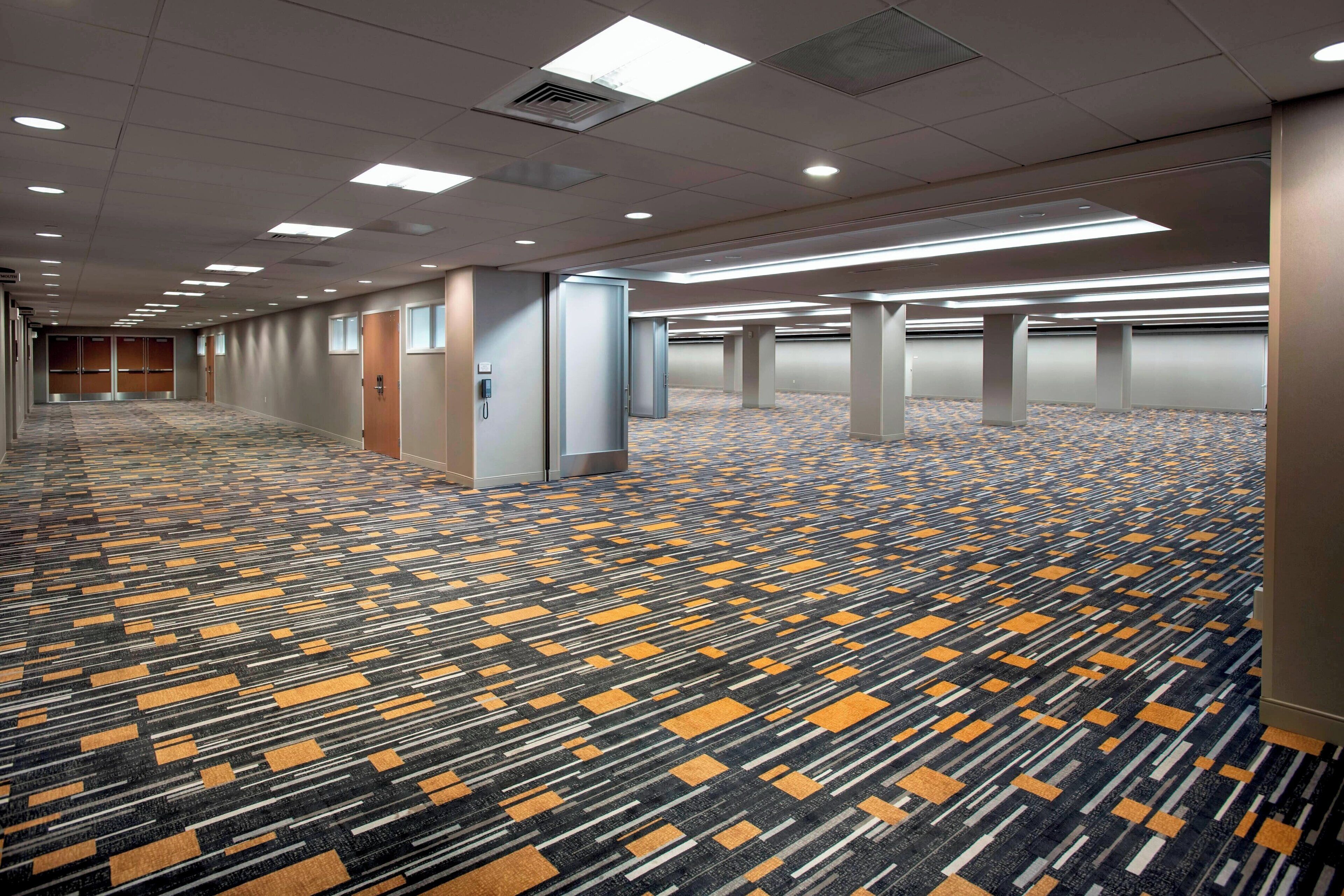 A long, narrow hotel corridor with a patterned carpet, recessed lighting, and a series of doors leading to individual rooms.