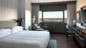 A hotel room with a large bed, a desk, a chair, and a view of the city through a large window.