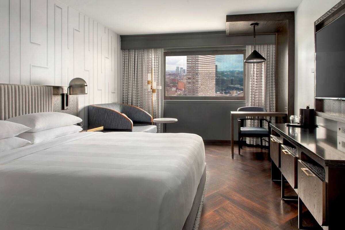A hotel room with a large bed, a desk, a chair, a TV, and a window with a city view.