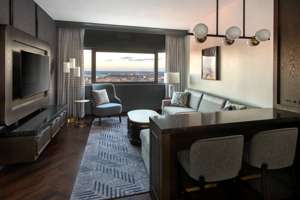A modern hotel room with a large TV, comfortable seating, a bar area, and a view of the city.