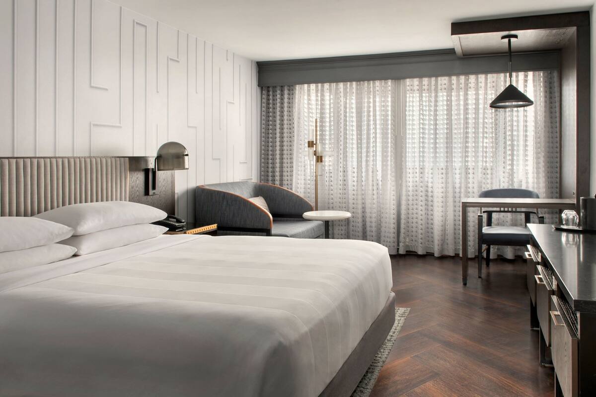 A modern hotel room with a large bed, a desk, and a chair. The room has a neutral color scheme with white and gray tones.
