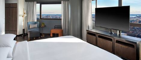 A hotel room with a large bed, a TV, a desk, and a view of the city.