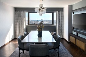 A modern dining area with a rectangular table, six chairs, a vase with flowers, a large window with curtains, and a flat-screen TV on the wall.