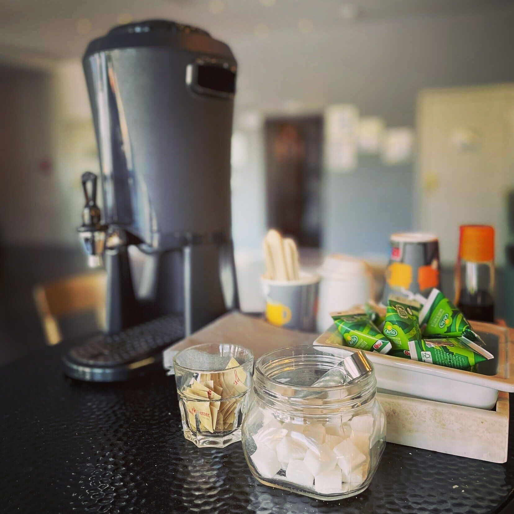Eine Küchenarbeitsplatte mit Kaffeemaschine, einem Glas mit Eiswürfeln, einer Schale mit Grünteebeuteln und einer Kaffeetasse.