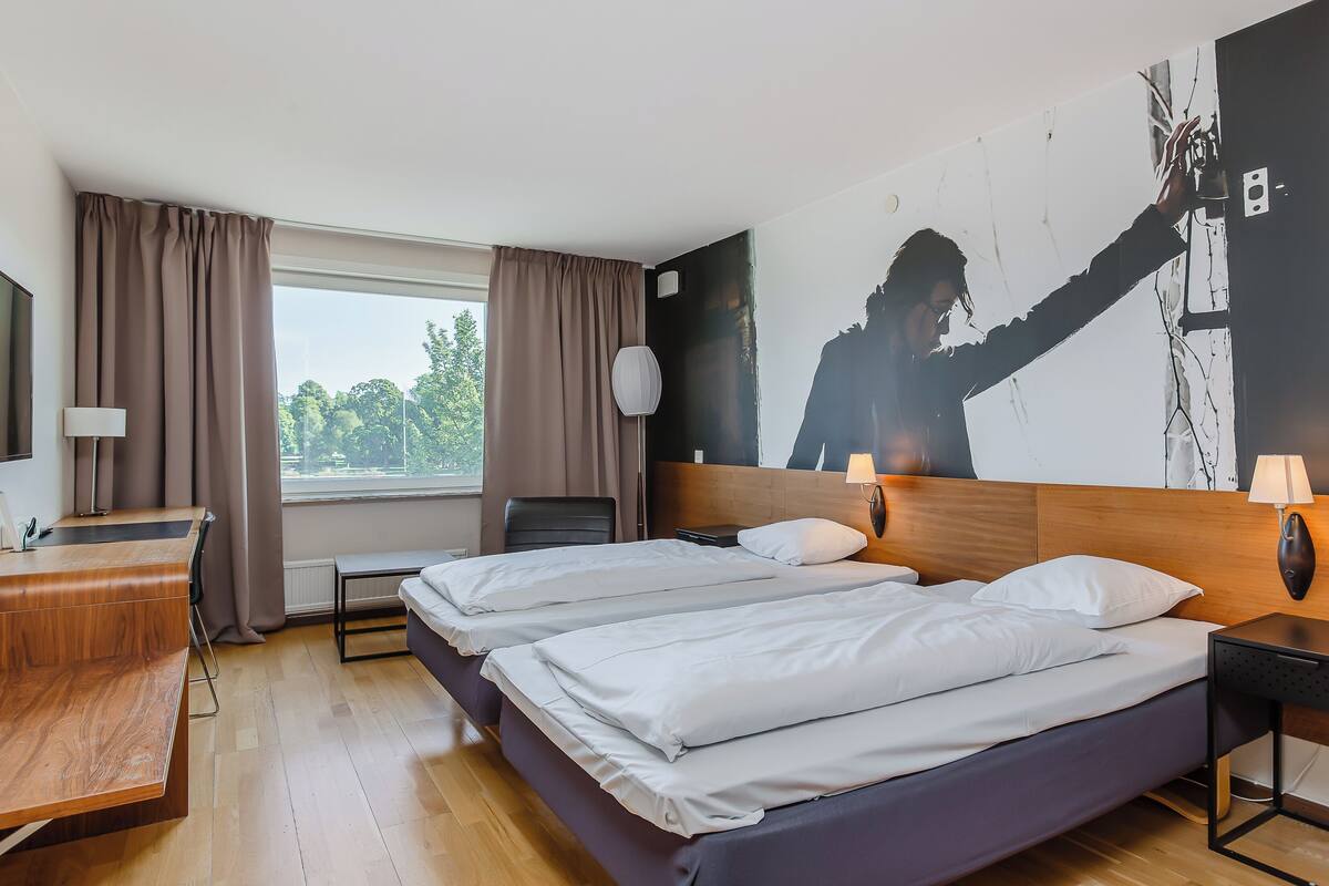Ein Hotelzimmer mit zwei Betten, einem Schreibtisch, einem Stuhl, einem Fenster mit Vorhängen und einem Wandbild.