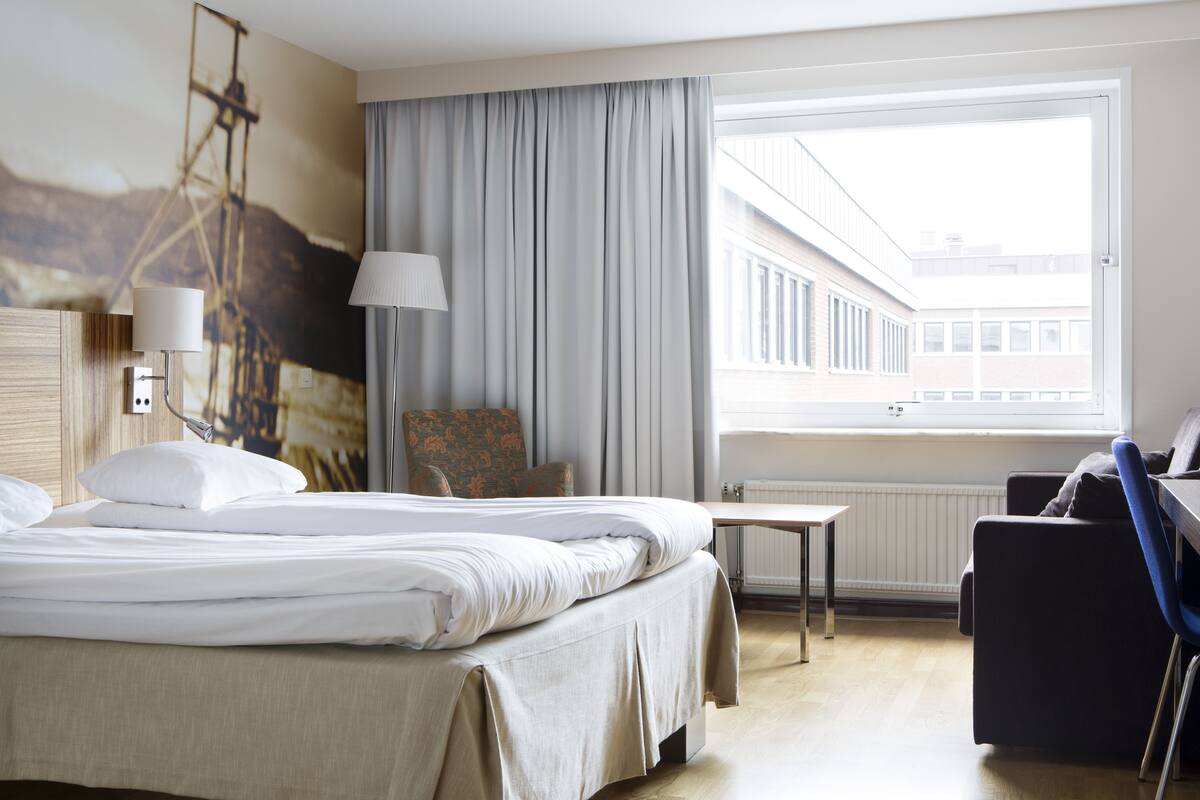 Ein Hotelzimmer mit Bett, Schreibtisch, Stuhl, Lampe, Fenster mit Blick und einem Bild an der Wand.