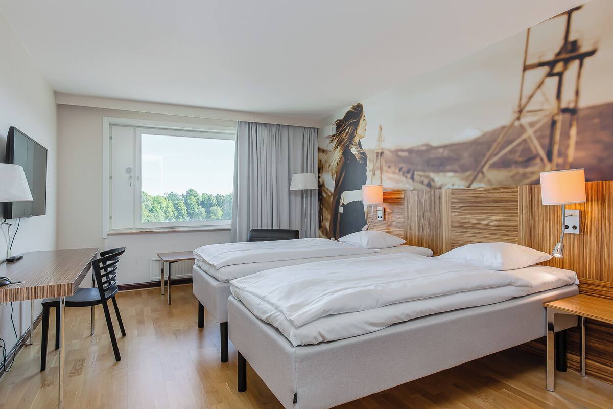 Ein Hotelzimmer mit zwei Betten, einem Schreibtisch, einem Stuhl, einem Fernseher und einem Gemälde einer Brücke.