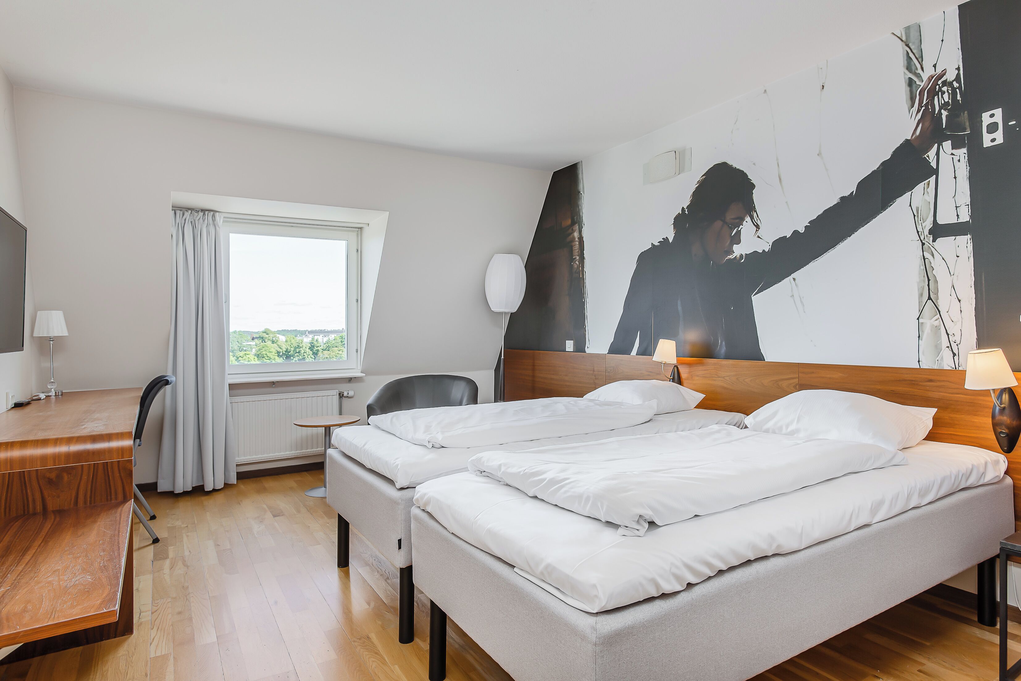 Habitación de hotel con dos camas, un escritorio, una silla, una ventana con cortinas y una pared con un cuadro abstracto grande.