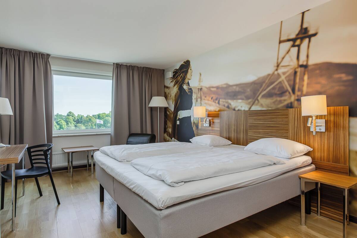 Ein Hotelzimmer mit einem Bett, zwei Nachttischen, einem Schreibtisch mit Stuhl und einem großen Fenster mit Vorhängen.