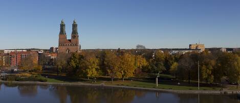 A historic building with two towers, a park with autumn trees, and a calm body of water reflecting the scene.