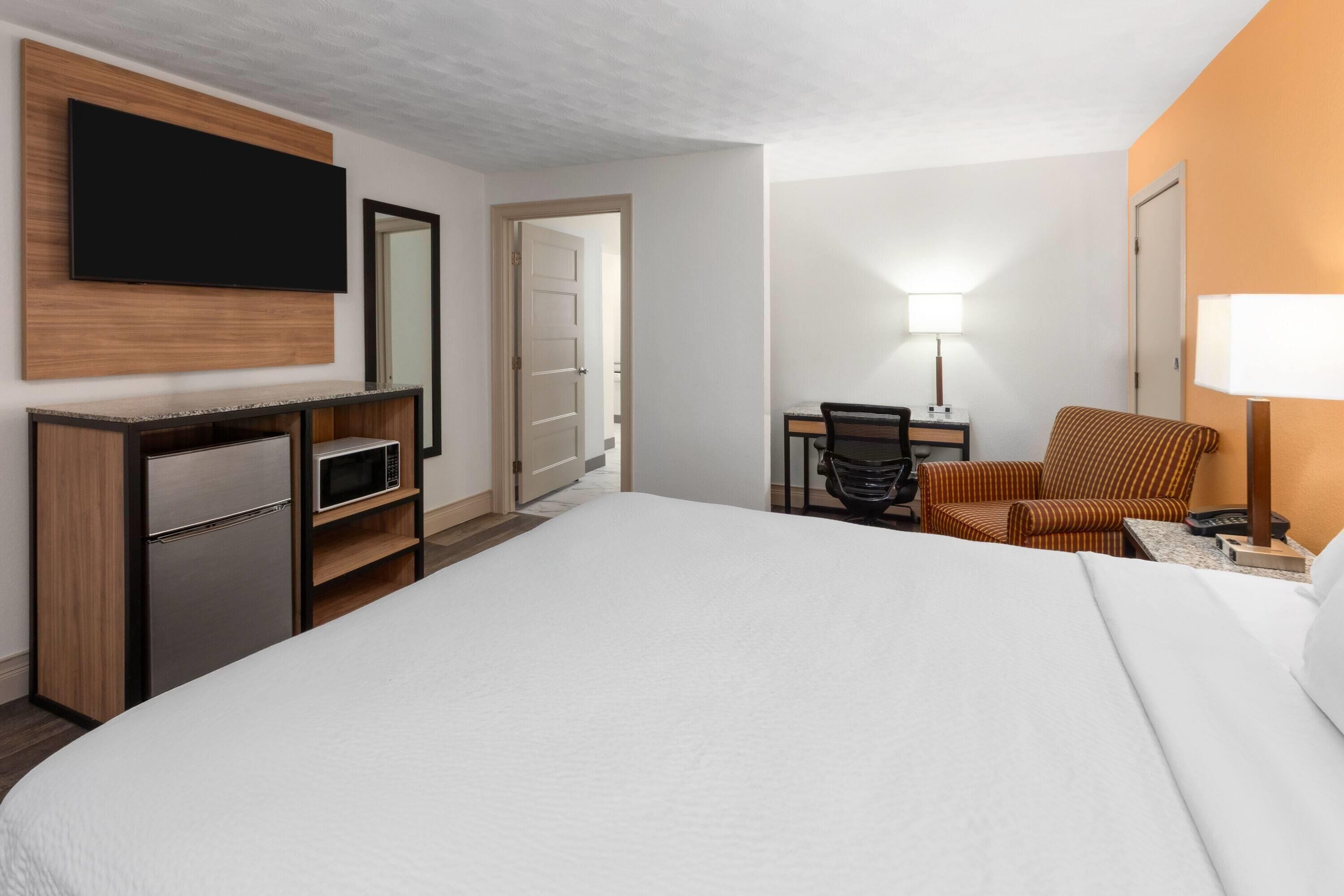 A hotel room with a bed, a desk with a chair, a television mounted on the wall, and a lamp on the desk.