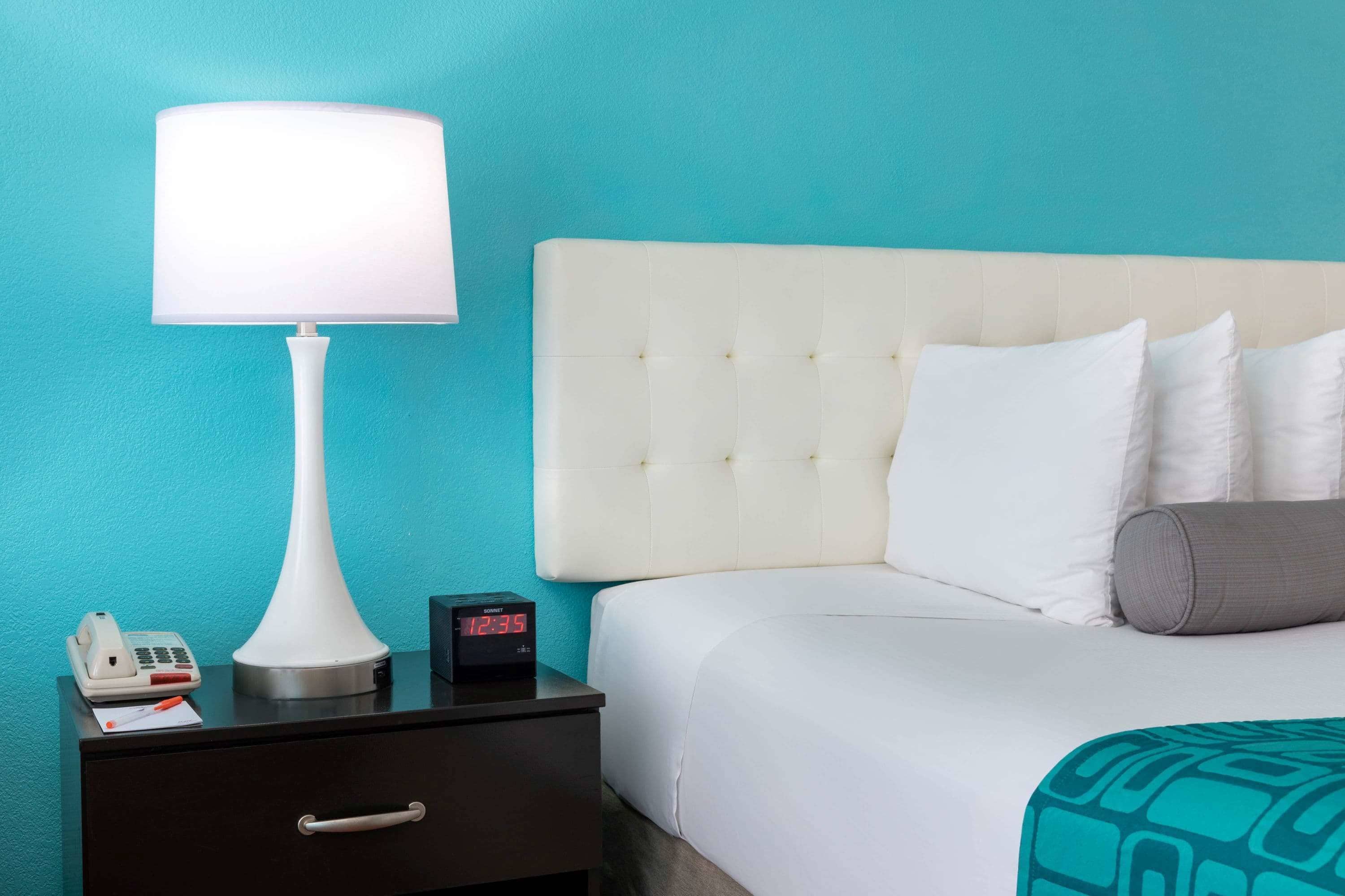 Een hotelkamer met een bed, nachtkastje, lamp, telefoon en digitale klok.