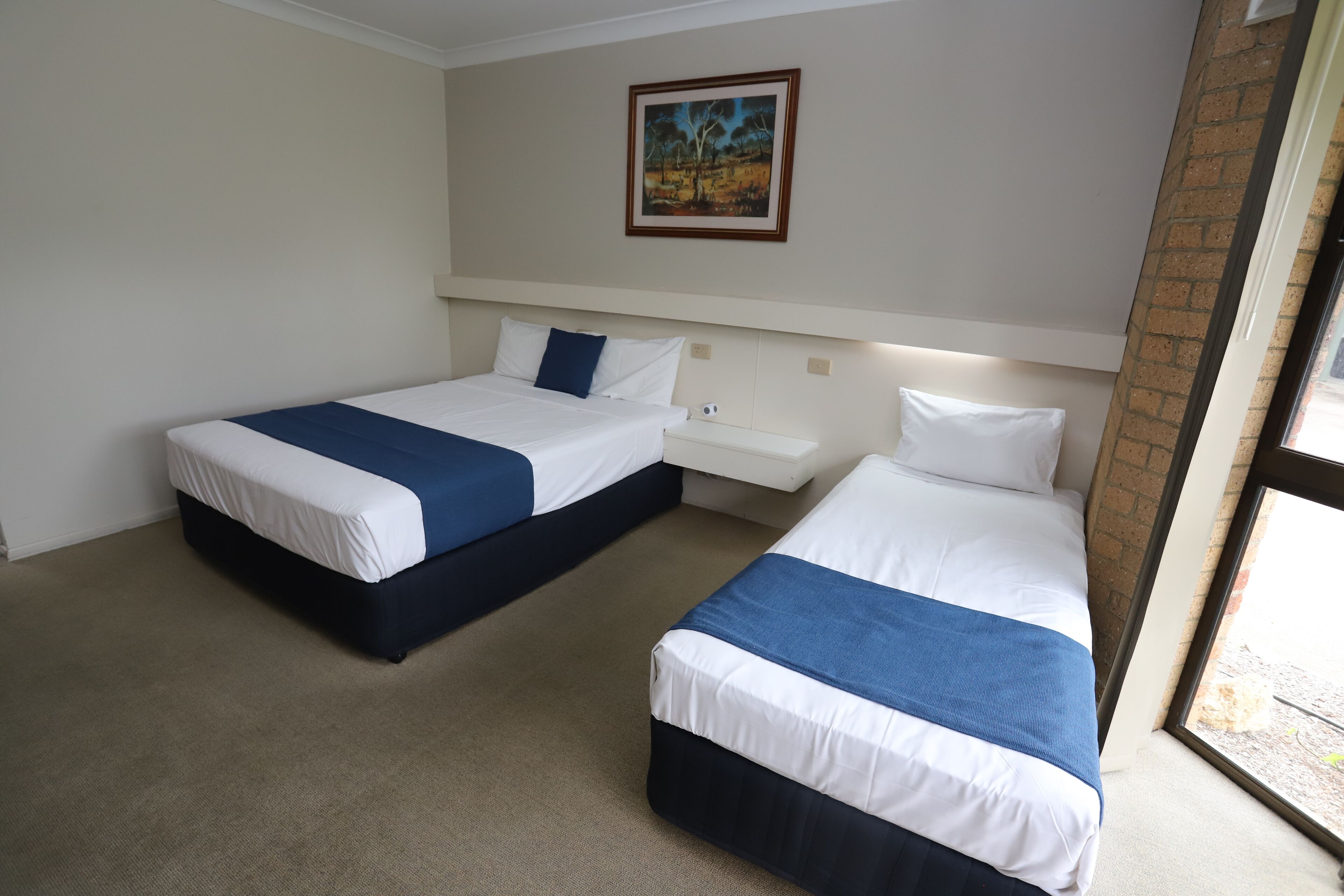 A hotel room with two single beds, a nightstand, a painting on the wall, and a window with a view.