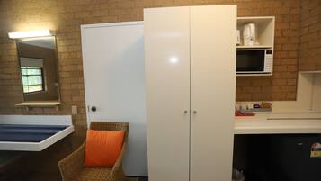 Una stanza con un muro in mattoni, una porta bianca, una sedia in vimini con cuscino arancione, un armadietto bianco e un forno a microonde sopra un lavello.