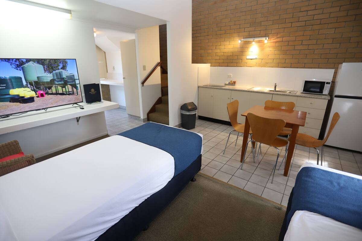 Quarto de hotel compacto com cama, escrivaninha com computador, pequena cozinha e mesa de jantar com cadeiras.