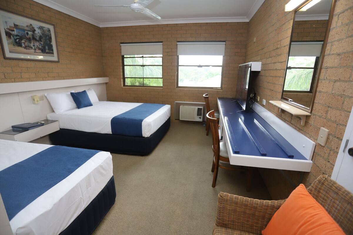 Quarto de hotel com duas camas, uma escrivaninha, uma cadeira, um espelho grande e uma janela.