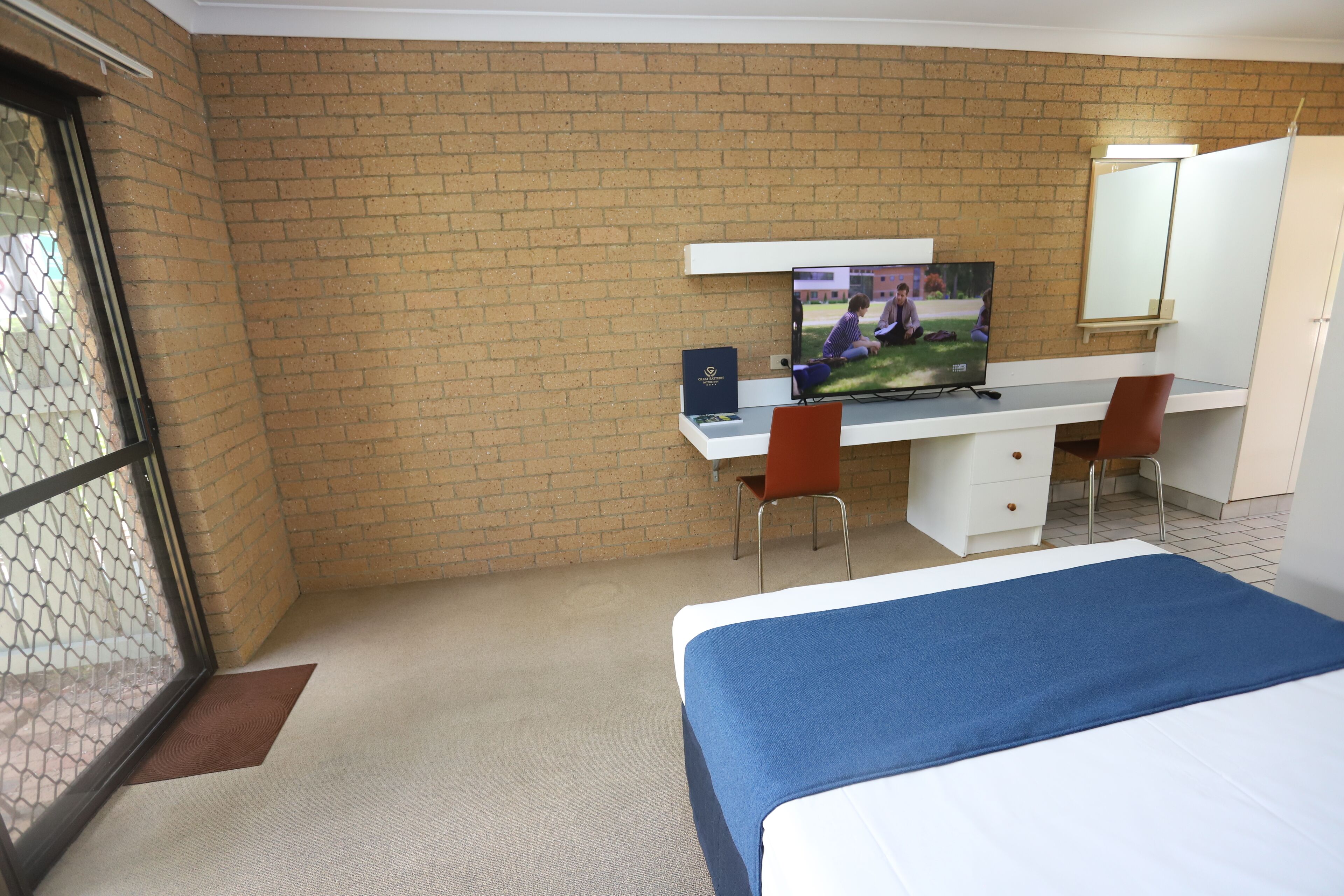 Quarto de hotel com parede de tijolos, uma TV, uma escrivaninha com cadeira e uma cama com colcha azul.