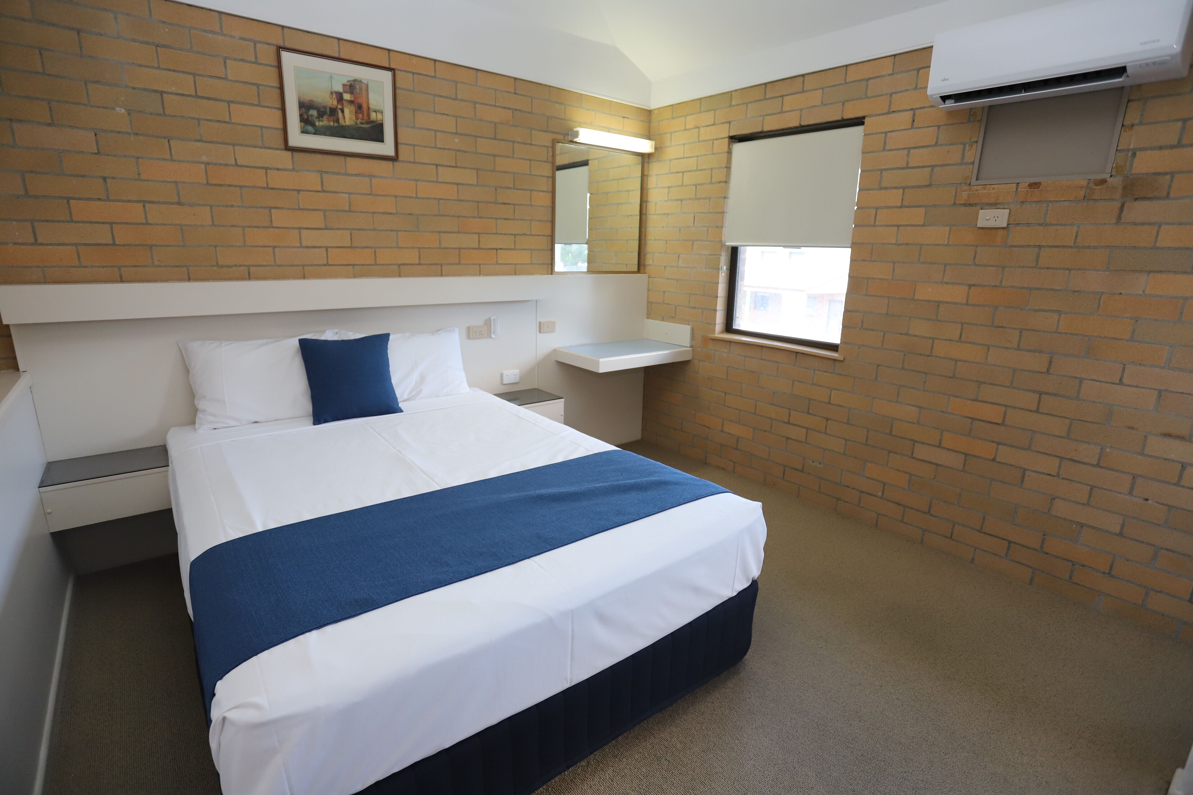 A hotel room with a bed, a small bedside table, a window with blinds, a wall-mounted air conditioner, and a picture on the wall.