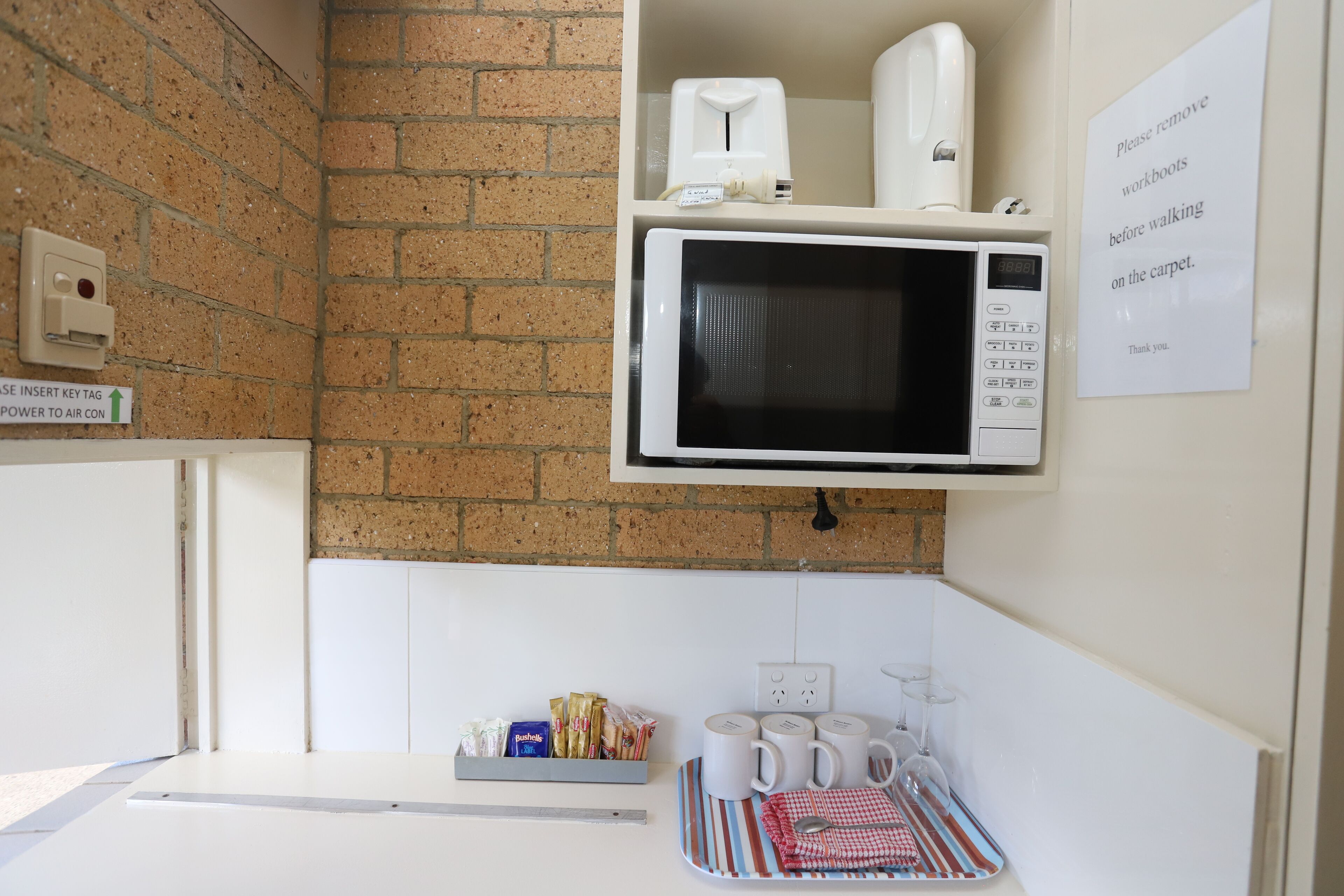Uma cozinha compacta com micro-ondas, torradeira e cafeteira. Há um aviso pedindo para retirar os cadernos antes de pisar no carpete.
