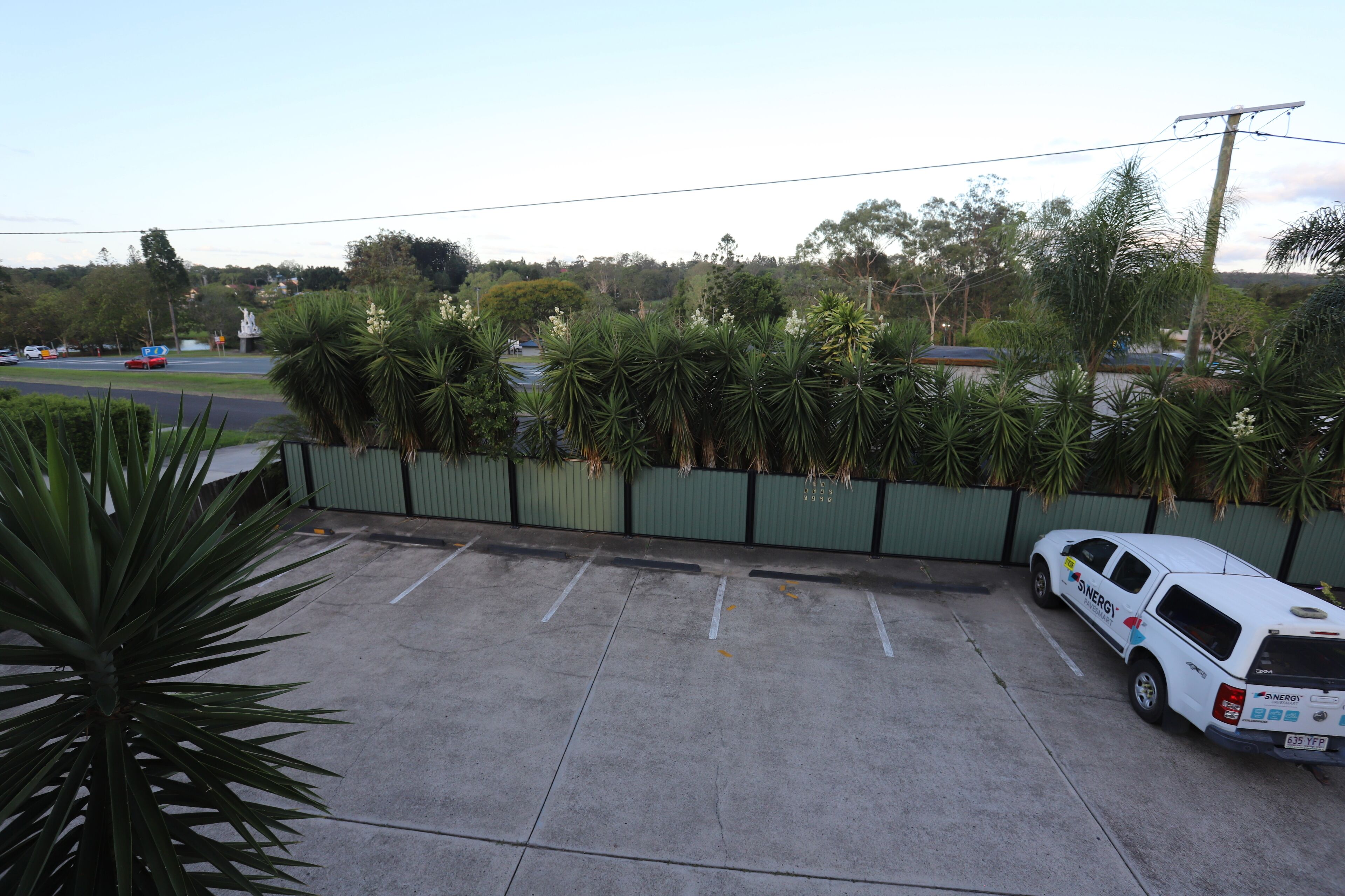 Un área de estacionamiento con un vehículo utilitario blanco, una cerca verde y una hilera de palmeras.