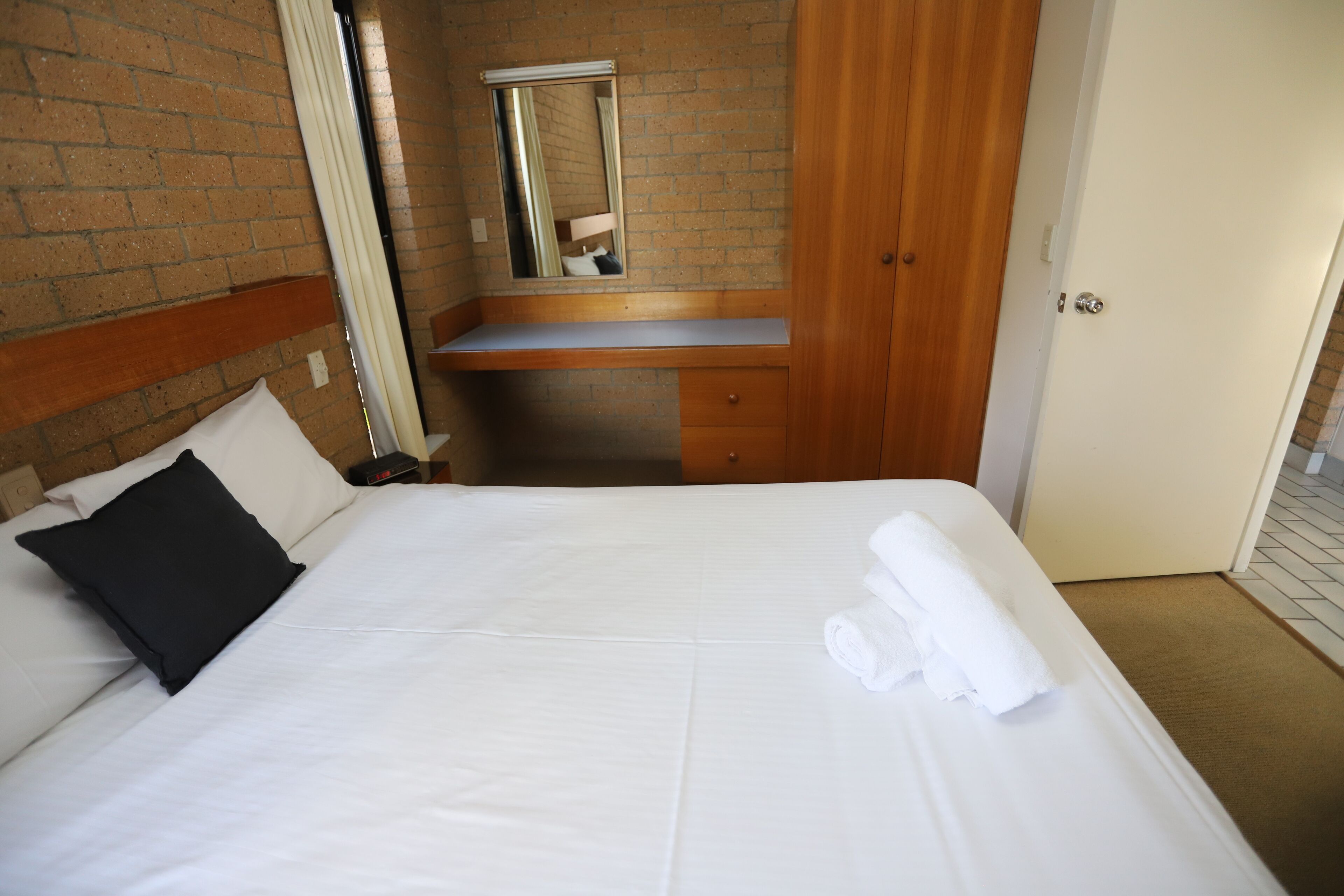 A hotel room with a bed, a wooden nightstand, a mirror, and a door.