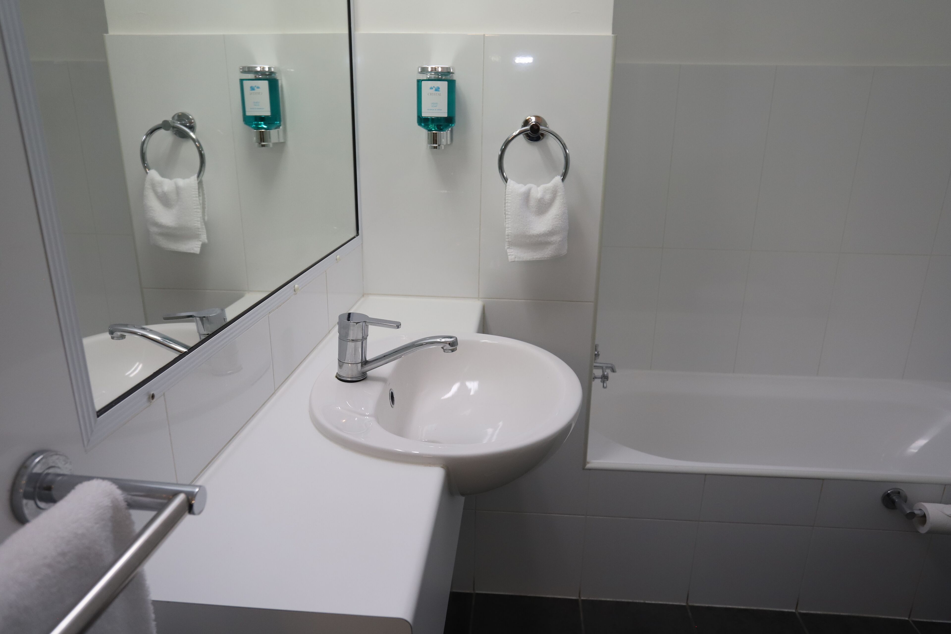 A bathroom with a white sink, a mirror, a towel rack, a bottle of soap, and a bathtub.