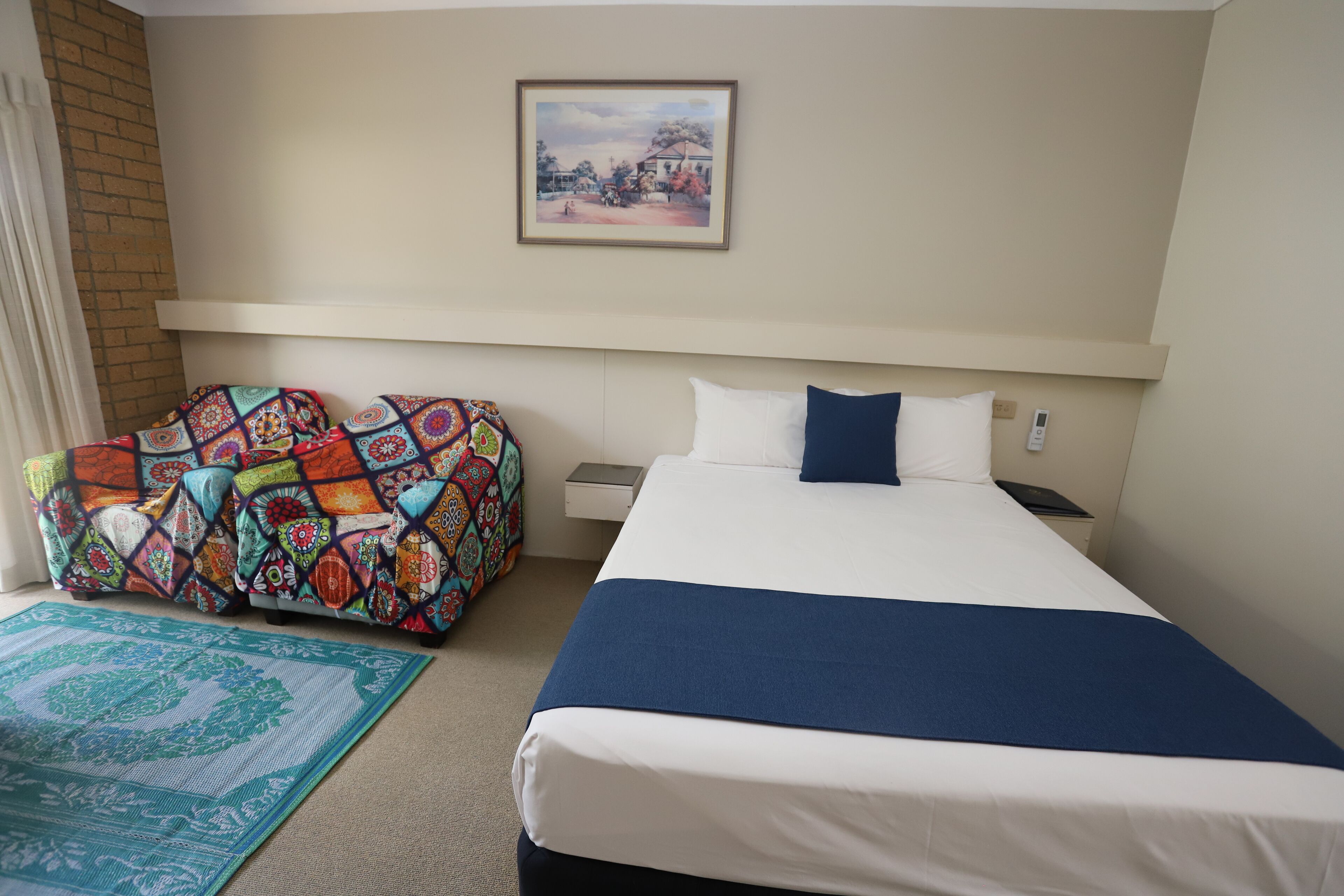 A hotel room with a large bed, a colorful sofa, a framed picture on the wall, and a small bedside table.