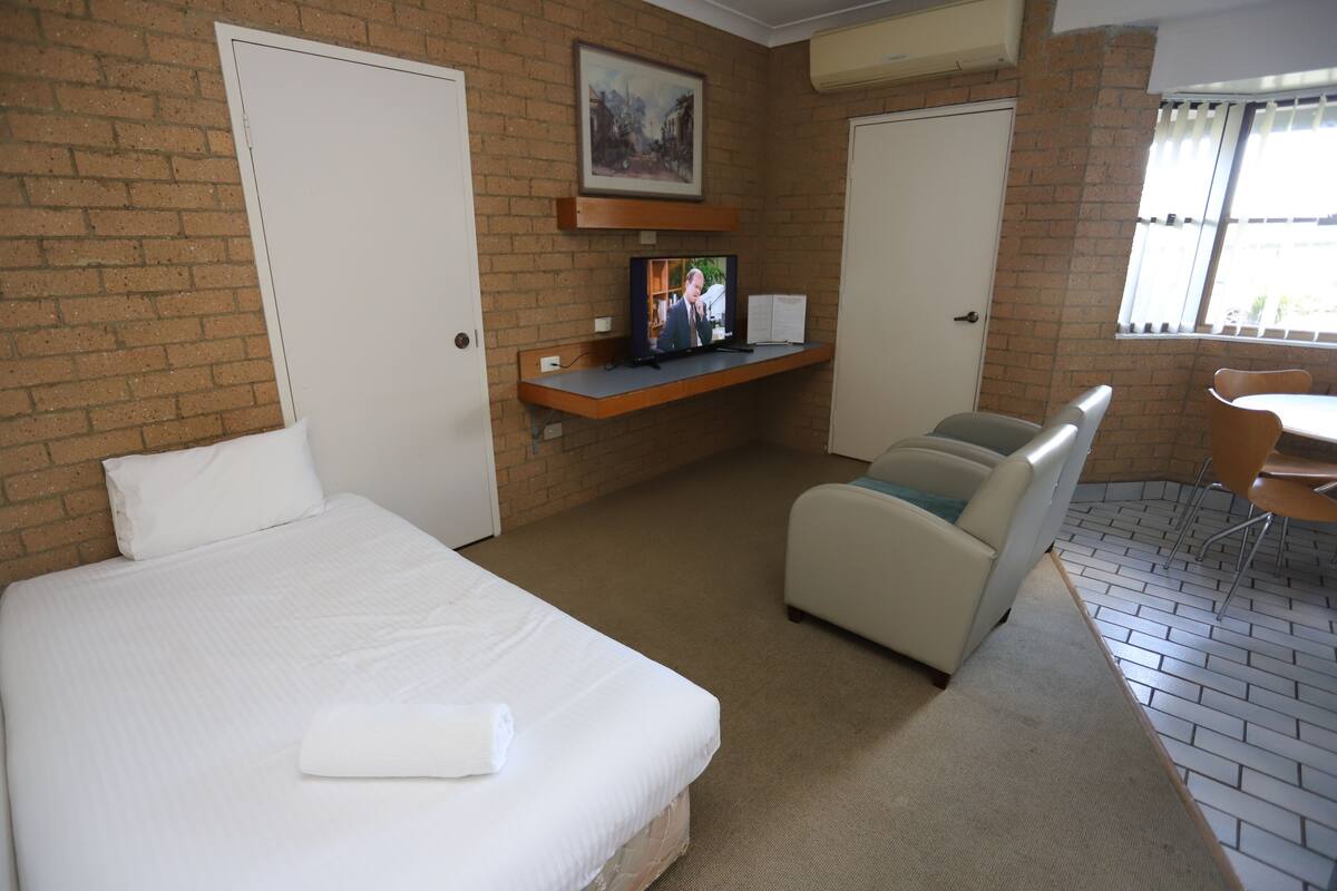 Quarto de hotel com cama, área de estar com cadeira e mesa, televisão em prateleira e janela com persianas.