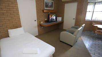 A hotel room with a bed, a seating area with a chair and a table, a television on a shelf, and a window with blinds.