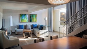 A modern living room with a sofa, coffee table, and dining table. There is a staircase with a railing and a large window with curtains. - Houston Airport Marriott at George Bush Intercontinental (Houston)