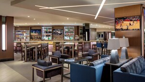 A modern bar area with seating, a television displaying a sports event, and a variety of bottles on shelves. - Houston Airport Marriott at George Bush Intercontinental (Houston)